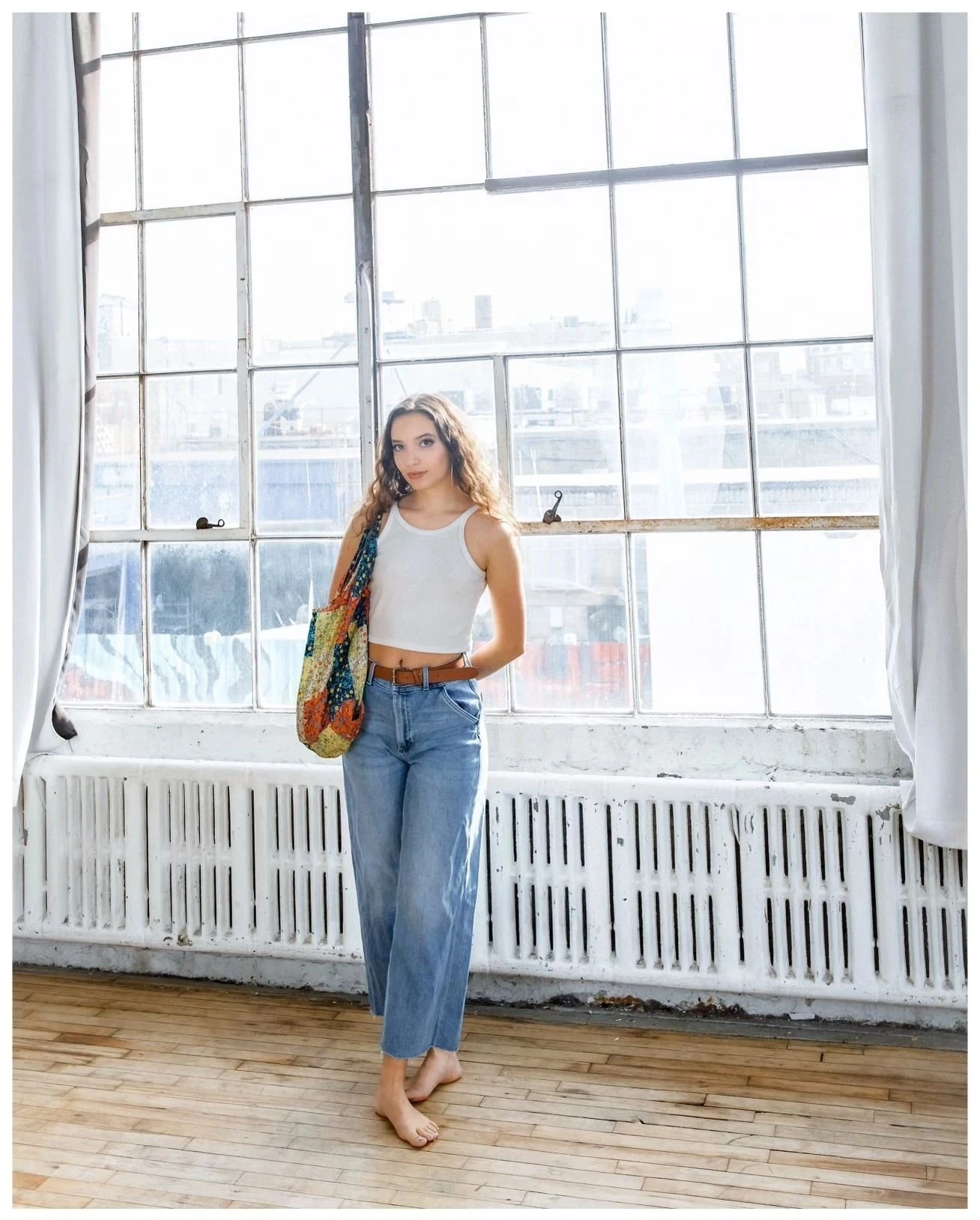 Lauren Maria Medina by a sunlit studio window, wearing a white top and jeans